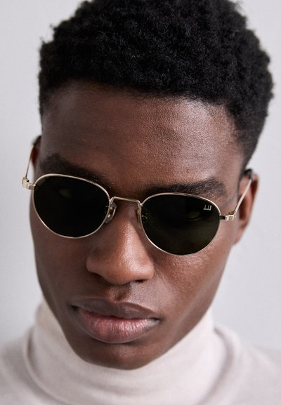 Round sunglasses with black lenses, thin gold metal frames, and adjustable temples. Model wears a light beige turtleneck sweater.