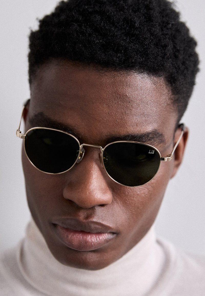 Pyöreät aurinkolasit mustilla linsseillä, ohuilla kultaisilla metallikehyksillä ja säädettävillä temppeleillä. Mallilla on päällään vaaleanbeige pooloneule.