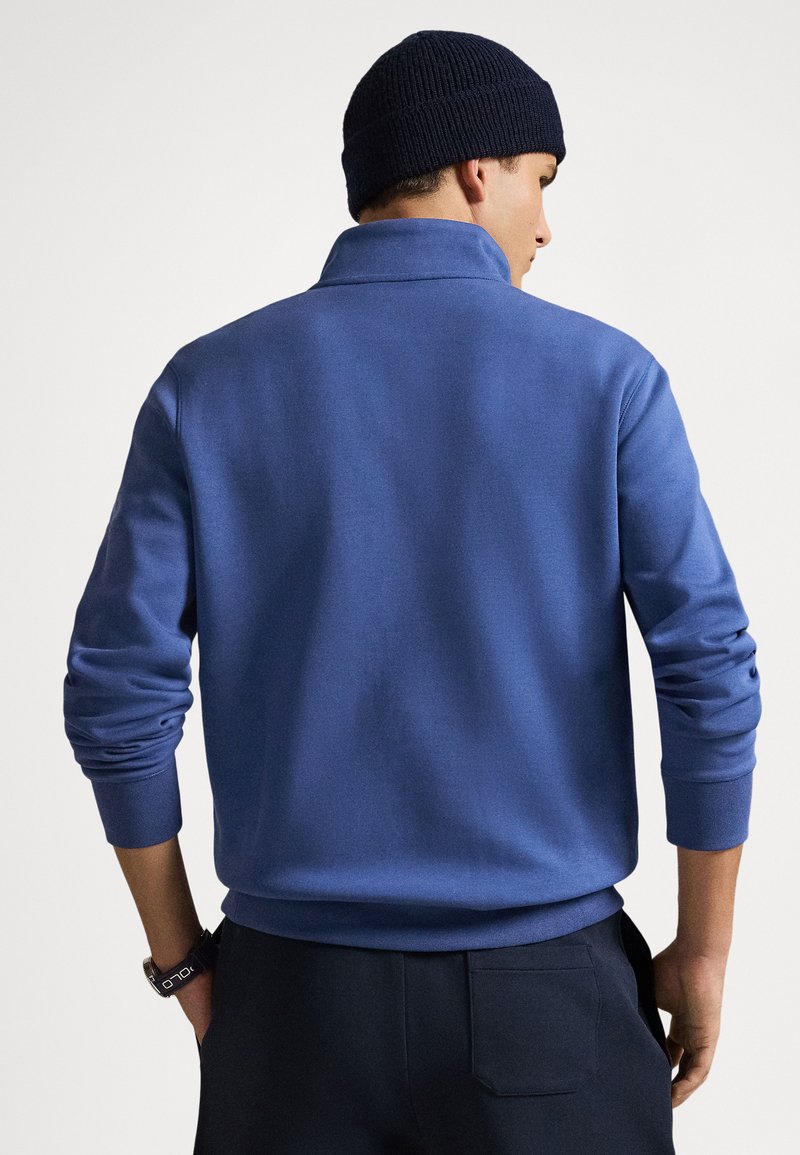 Uomo con cuffia blu scuro, maglione a maniche lunghe blu e pantaloni neri, in piedi con le mani nelle tasche, rivolto di spalle.