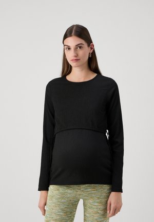 Svart langermet topp med teksturert stoff, rund hals og elastisk midje designet for graviditet. Kombinert med mønstrede grønne bukser.