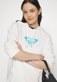 White hoodie made of soft fabric, featuring a turquoise heart and diamond logo print. Includes drawstrings and ribbed cuffs.