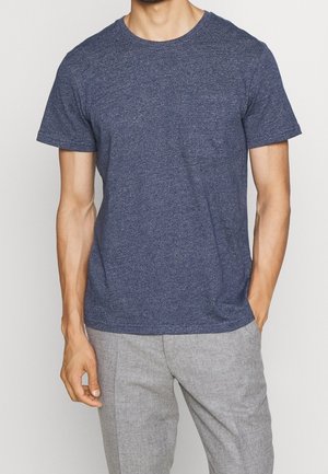 Granatowy t-shirt z krótkim rękawem wykonany z teksturowanej tkaniny, z okrągłym dekoltem i małą kieszonką na piersi. Noszony z jasnoszarymi spodniami.