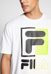 Biała koszulka z krótkim rękawem z dużym logo FILA w czerni i jasnej zieleni. Koszulka ma okrągły dekolt i gładką strukturę.
