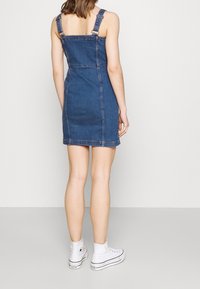 Kvinna som står vänd bort ifrån kameran, klädd i en ärmlös denimminiklänning och vita högstövliga sneakers, framför en enfärgad bakgrund.