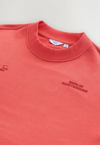 Sudadera de color coral con un cuello de canalé; presenta "BERLÍN" y "ÁMSTERDAM" en letras de color rojo oscuro. Textura suave, tejido de peso medio.