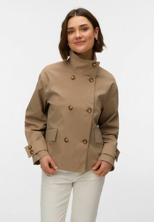 Kvinde med kort brunt hår iført en lysebrun dobbeltradet jakke og hvide bukser, smilende med hænder tæt på jakkeens kant.