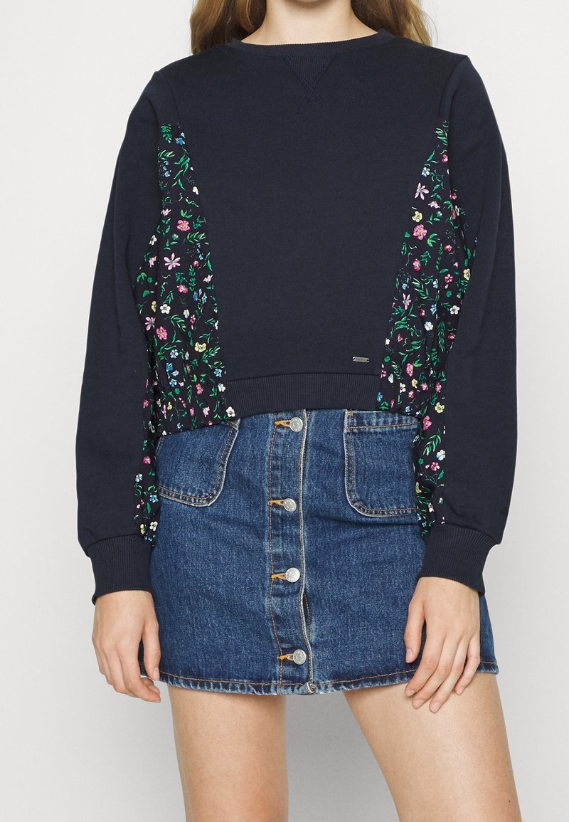 Femme portant un sweat-shirt bleu marine à panneaux latéraux fleuris et une mini-jupe en jean bleue à boutons devant, debout sur un fond uni.