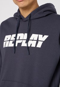 Marineblaues Hoodie mit großem weißen "REPLAY"-Druck auf der Vorderseite, mit Kapuze und Kordelzug sowie gerippten Bündchen. Weicher Stoff.