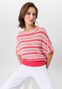 Rood-witte top met een chevronpatroon en een geribbeld koraalkleurig elastiek aan de taille. Korte mouwen en een bootnek. Gekocht met een witte pantalon.