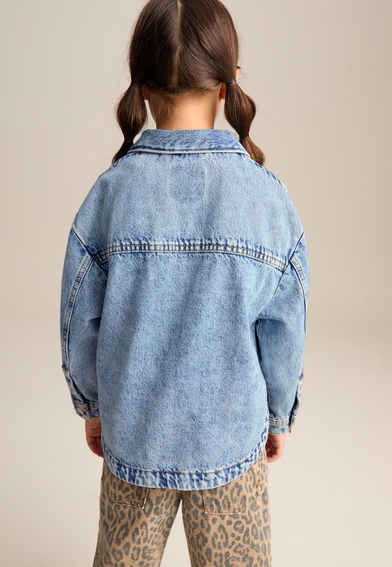 Kind met bruin haar in vlechtjes, draagt een lichtblauwe spijkerjack en broek met luipaardprint, met de rug naar de camera toe.