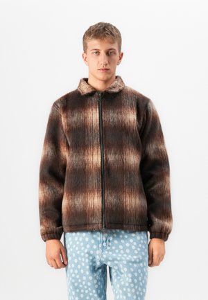 Jonge man in een bruin en beige geruite fuzzy zip-up jack en lichtblauwe jeans met witte stippen, staand voor een witte achtergrond.