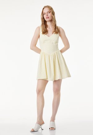 Jonge vrouw met lang haar, gekleed in een mouwloze gele ooglet-jurk en witte open-teen hakmuiltjes, staand met de handen achter de rug.