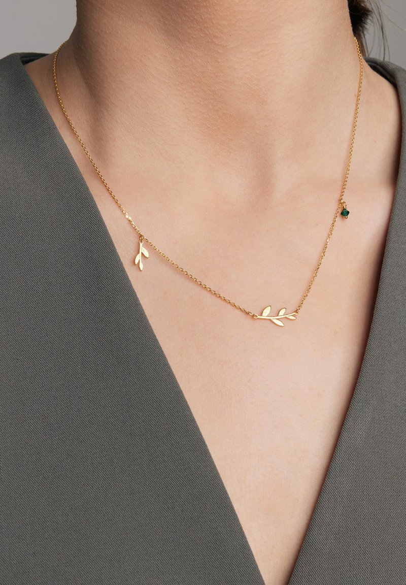 Złoty naszyjnik z delikatnym wzorem gałązki i dwoma akcentami liści, zawieszony na cienkim łańcuszku, na tle szarej tkaniny w serek.