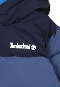 Giacca isolante blu e blu navy con design imbottito, dotata di logo bianco "Timberland" e emblema dell'albero. Tessuto liscio, leggermente lucido.