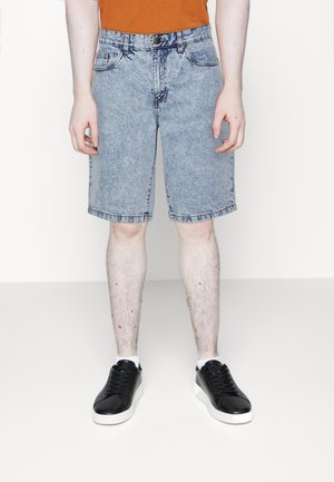 Personne portant un short en jean délavé à l'acide, des baskets basses noires, des chaussettes blanches et un haut orange, debout devant un fond uni clair.