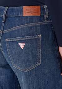 Jeans in denim blu scuro con un'etichetta in pelle marrone con il logo "Guess Jeans" in rilievo. La tasca posteriore presenta un piccolo logo triangolare con la scritta "Guess".