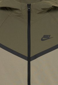 Priekinis vaizdas olivinės ir smėlio spalvos Nike striukės su juodu užtrauktuku ir logotipu ant krūtinės, pasižyminčios chevrono dizainu viršutinėje liemens dalyje.