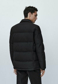 Giubbotto puffa nero con un design a quadrati, cuciture visibili e colletto. Il materiale appare liscio e lucido.