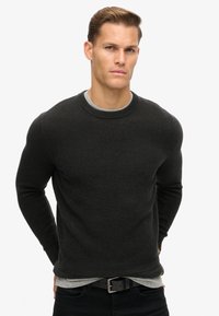 Svart strukturerad stickad tröja med rund halsringning och långa ärmar, bärs över en grå t-shirt med rund halsringning, kombinerad med svarta jeans och ett svart bälte.