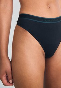 Melnas kokvilnas string biksītes ar kontrastējošu zilu jostu, uz kuras ir "Calvin Klein" logs. Gluda tekstūra, minimālistisks dizains un zemas vidukļa griezums.