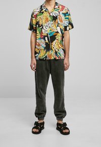 Chemise noire à manches courtes avec imprimé coloré de bananes et de feuilles, associée à un pantalon de jogging gris foncé et des sandales noires à brides.