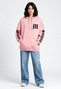 Felpa rosa chiaro con tasca a marsupio, testo grafico nero sulle maniche e sul petto, abbinata a jeans larghi azzurri.