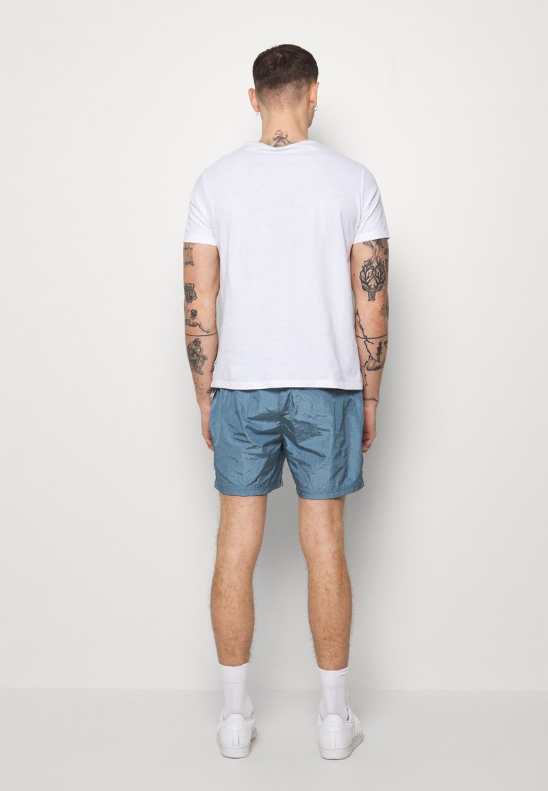 T-shirt blanc à col rond, manches courtes, associé à un short bleu clair, brillant et texturé. Le modèle porte des chaussettes blanches et des baskets.
