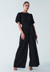 Zwarte jumpsuit met pofmouwen, een vierkante halslijn en een ceintuur die in een strik wordt geknoopt. Heeft wijde pijpen en een gladde, gestructureerde stof.