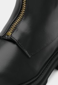 Botín de cuero negro con una cremallera dorada a lo largo del costado, con una textura suave y costuras reforzadas en el panel superior.