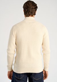 Kremowy sweter dziergany o fakturowanym wzorze diamentów, z prążkowanymi ściągaczami na mankietach i u dołu oraz wysokim kołnierzem. Noszony z ciemnymi jeansami.