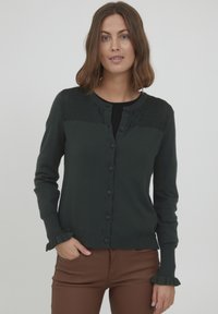 Tamnozeleni kardigan s teksturiranim jarmom, prednjim zatvaranjem na dugmad i volanima na manžetama. Izrađen od mekog materijala, ima uski dizajn.