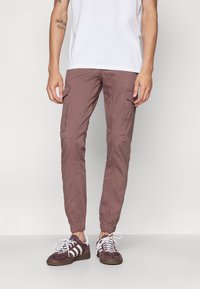 Muž oblečený v mauve cargo jogger kalhotách se elastickými manžetami a vínovo-bílých teniskách, stojící na pozadí s jednoduchým světlým pozadím.