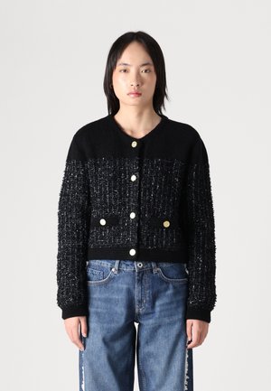 Svart cardigan med teksturert stoff, med gullknapper, to frontlommer og ribbede mansjetter. Bært sammen med blå, løstsittende jeans.