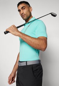 Polo tričko s krátkym rukávom v tyrkysovej farbe s tromi gombíkmi a logom, kombinované so sivým opaskom a čiernymi nohavicami, drží golfovú palicu cez plece.