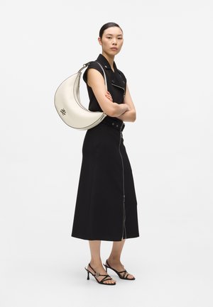 Ženska v črni obleki brez rokavov in sandalih nosi veliko belo torbo v obliki polmeseca čez ramo ter stoji s prekrižanimi rokami.