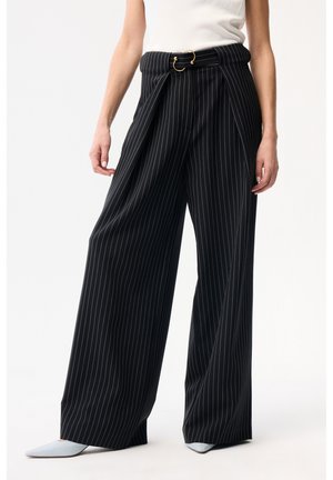 Frau trägt schwarze, weit geschnittene Nadelstreifenhosen mit passendem Gürtel und weißen spitzen Schuhen, steht vor einem schlichten Hintergrund.