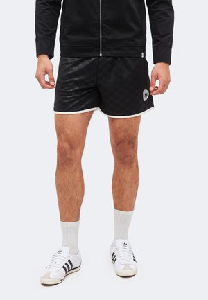 Mies, joka käyttää mustia ruudullisia urheilushortseja, joissa on valkoinen reunus, valkoiset sukat ja valkoiset Adidas-tossut mustine raitoineen.