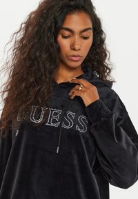 Felpa nera in velluto con cappuccio regolabile e decorazioni in strass che formano la scritta "GUESS" sul davanti. Texture morbida e vestibilità rilassata.