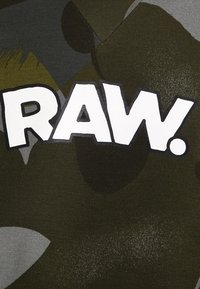 T-shirt camouflage vert avec un grand logo en texte blanc "RAW." entouré de noir. Le matériau semble être du coton avec une texture lisse.