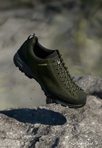 Dunkelgrüner Wanderschuh mit einem Obermaterial aus Wildleder, verstärkter Zehenkappe, langlebiger Gummisohle und grünen Akzent-Schnürsenkeln, der über einer felsigen Oberfläche schwebt.