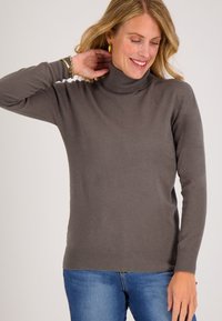 Grijze coltrui met een zachte textuur, met lange mouwen en een geribbelde zoom. Gecombineerd met blauwe jeans, tegen een effen achtergrond.