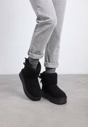 Botas de ante negras hasta el tobillo con un puño texturizado y plush, y una suela de goma plana. Combinadas con jeans grises doblados, contra un fondo neutro.