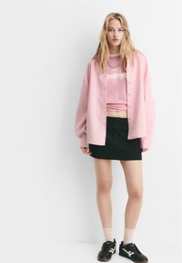 Giovane donna che indossa una camicia a righe rosa con bottoni sopra una t-shirt grafica rosa, una gonna nera, calze chiare e sneakers nere, in piedi contro un muro bianco.