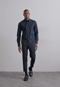 Mörkblå skjorta med knäppning och långa ärmar, matchad med mörka byxor. Modellen bär svarta loafers och strumpor, mot en enfärgad bakgrund.