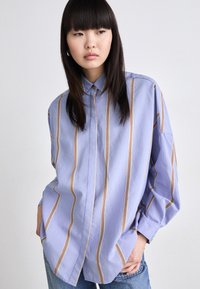 Light blue collared shirt with vertical stripes in orange and white. Loose fit with long sleeves, featuring a curved hem. Casual design.