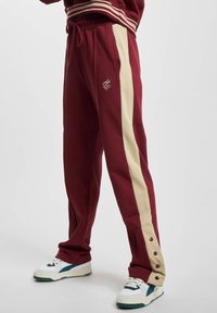 Mørkerøde sweatpants med cremefarvede sidelister, justerbar talje og trykknapdetaljer ved anklerne. Broderet logo på venstre ben.