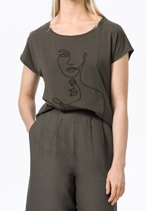 Femme portant un t-shirt vert olive foncé avec un dessin au trait noir de deux visages abstraits, associé à un pantalon taille haute assorti.