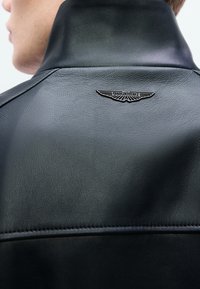 Fekete bőrkabát magas gallérral és ívelt varrási részletekkel, a hátán ezüst Aston Martin logóval díszítve.