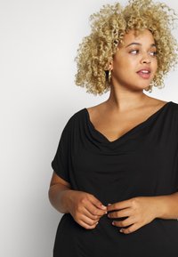 Sort draperet top med en bred halsudskæring og korte ærmer, lavet af et glat stof. Modellen har krøllet blond hår og hoopsøreringe.