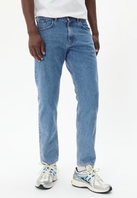 Jean en denim bleu clair avec une coupe décontractée, des coutures visibles, cinq poches et une taille classique, assorti avec des chaussures de sport argentées et bleues.